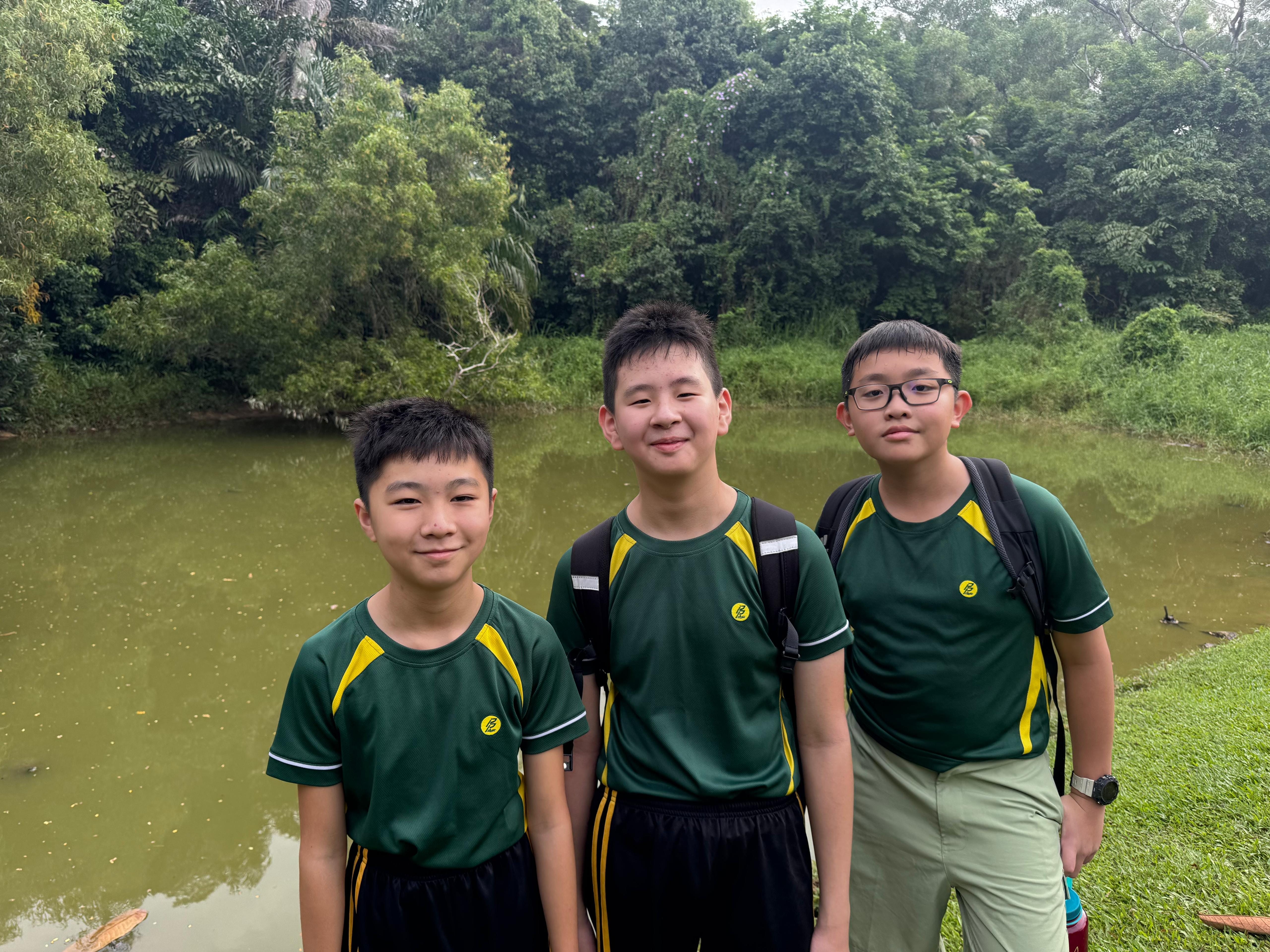 Photograph of three boys wearing matching green and yellow sports shirts standing near a pond with dense trees in the background. Two boys wear black shorts, one wears light-colored pants, and all have backpacks, suggesting an outdoor school or sports activity.